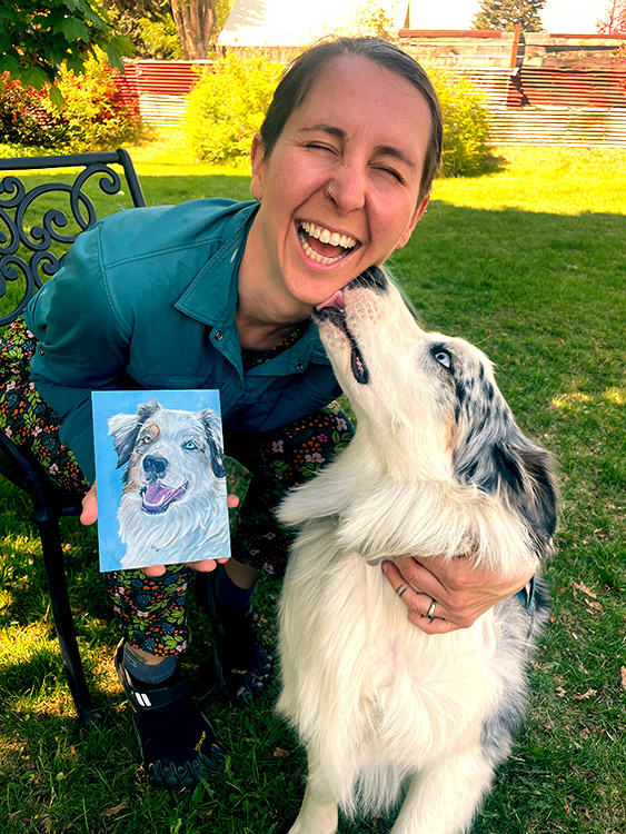 Artist's face is licked by dog as she holds painting of same dog.